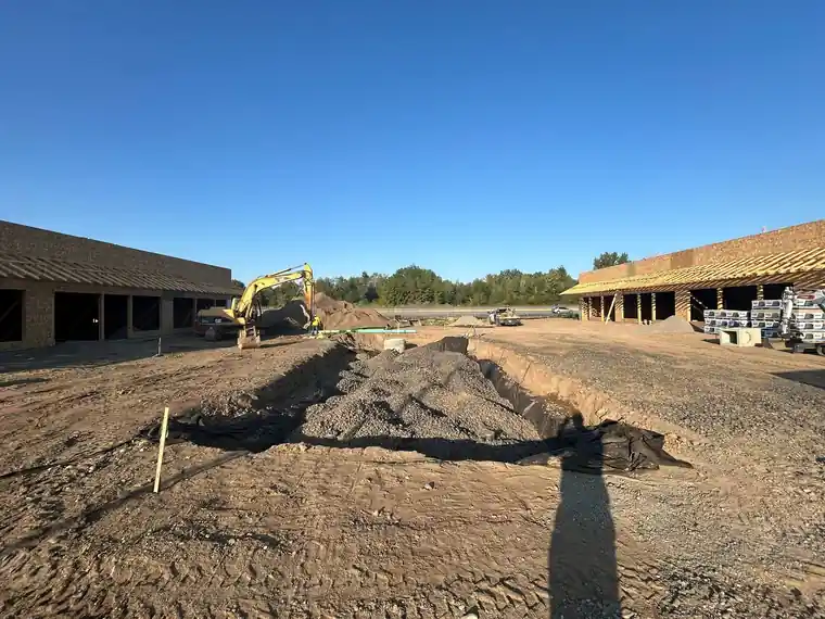 Open excavation between two building shells, with an excavator working.