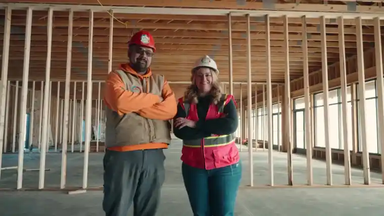Two crew members in hard hats inside a stud-framed building.
