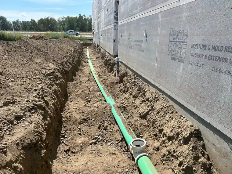 A trench with green sewer pipe along a wrapped building wall.